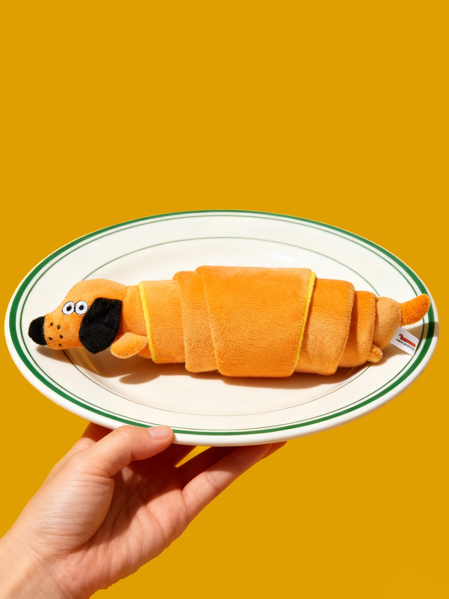 Dog toy shaped like a croissant on a white plate with a green rim against a yellow background